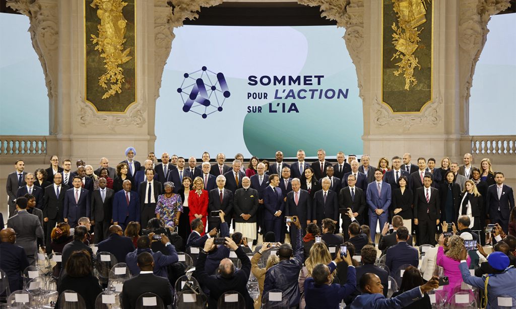 A wide group photograph from the AI Action Summit in Paris, featuring world leaders including Prime Minister Narendra Modi and French President Emmanuel Macron, alongside delegates from Saudi Arabia and other nations. They are posed together on a stage at the Grand Palais, symbolizing a unified global approach to AI governance and ethics.